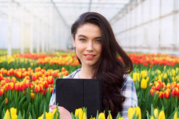 Uma jovem florista sorridente está de pé com um tablet nas mãos em uma estufa