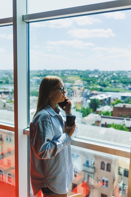 Uma jovem freelancer de óculos fica com café e fala ao telefone perto de uma grande janela e olha para a cidade Uma garota de camisa e shorts fica em um café perto da janela