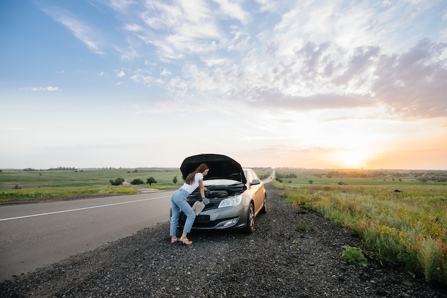 Uma jovem frustrada fica perto de um carro quebrado no meio da rodovia durante o pôr do sol. Avaria e reparação do carro. Esperando por ajuda.