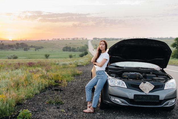 Uma jovem frustrada fica perto de um carro quebrado no meio da rodovia durante o pôr do sol. Avaria e reparação do carro. Esperando por ajuda.