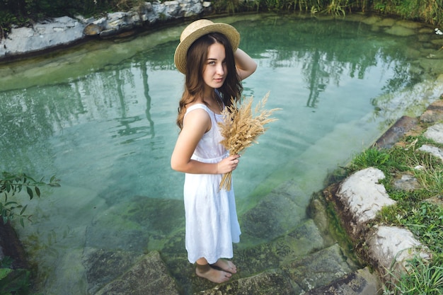 Uma jovem garota com um vestido branco em um dia ensolarado entra no lago. Menina com um chapéu de palha no rio ao pôr do sol.