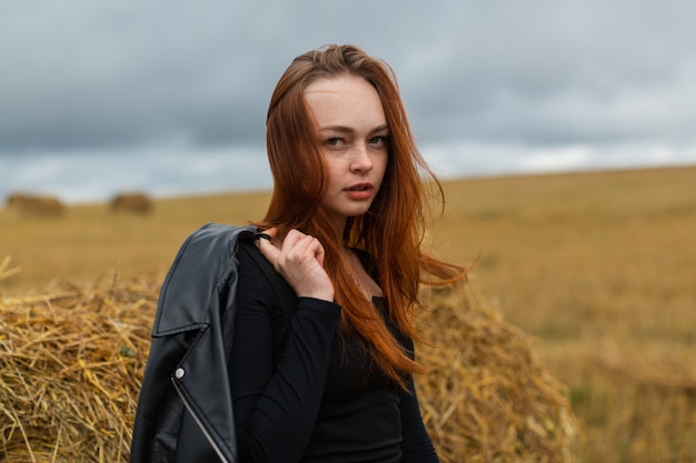 uma jovem garota de cabelo vermelho com uma jaqueta preta fica perto de um palheiro em um campo em tempo nublado. Caminhando na natureza em um dia de outono. Foto de alta qualidade