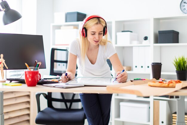 Uma jovem garota em fones de ouvido fica perto da mesa e tem um marcador na mão. Em cima da mesa está um quadro magnético.