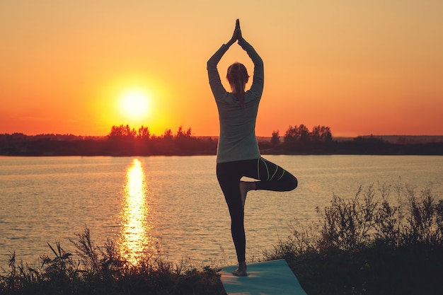 Uma jovem garota está de pé no lago ao pôr do sol, fazendo yoga.