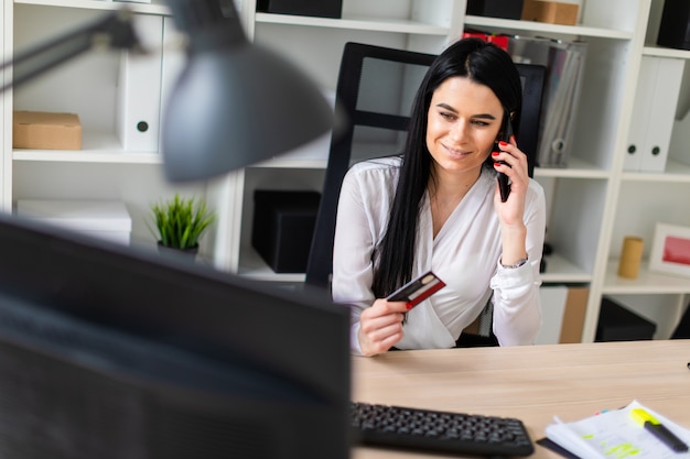 Uma jovem garota está sentada em uma mesa, segurando um cartão de banco na mão e falando ao telefone.