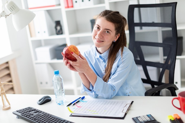 Uma jovem garota se senta em uma mesa no escritório e tem uma maçã na mão.