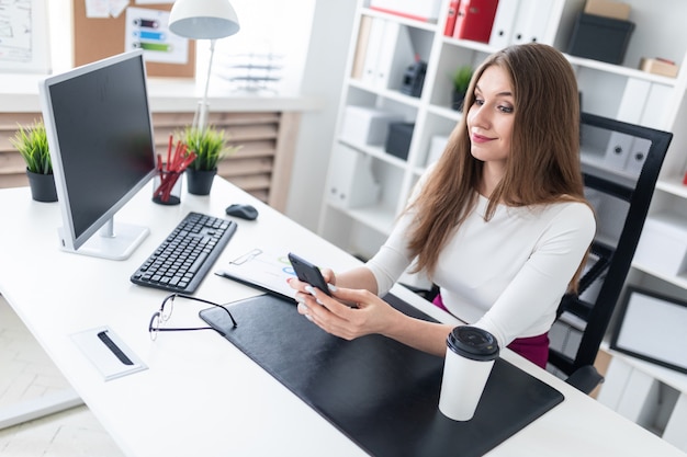 Uma jovem garota sentada em uma mesa no escritório e segurando um telefone. ao lado da mesa há um copo de café branco.