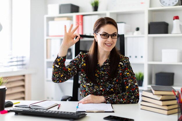 Uma jovem garota sentada no escritório à mesa do computador e mostra o sinal OK.