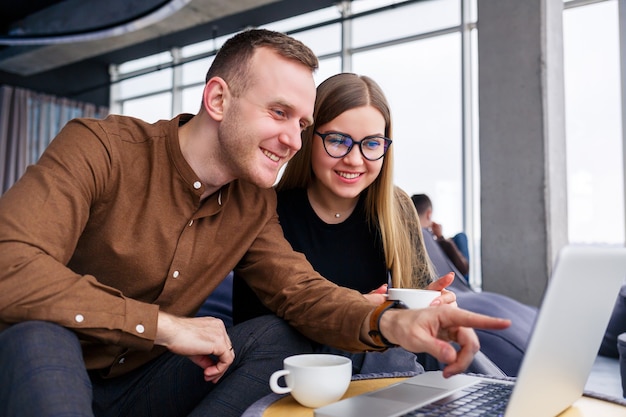 Uma jovem gerente de sucesso com seu chefe senta-se com um laptop em uma poltrona estofada perto da janela panorâmica e bebe café. Mulher de negócios e homem trabalhando em um novo projeto