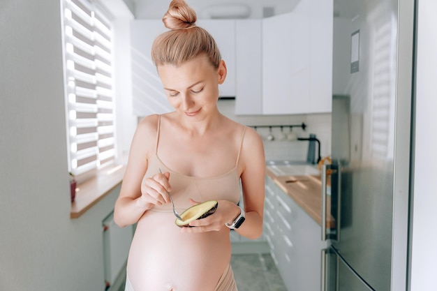 Uma jovem grávida de aparência europeia em sua cozinha com um abacate nas mãos o conceito de nutrição adequada de mulheres grávidas para o correto desenvolvimento do feto
