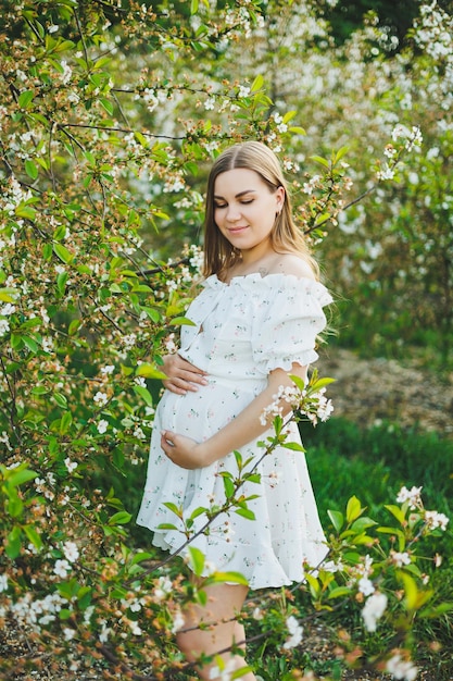 Uma jovem grávida em um vestido branco caminha em um pomar de maçã em flor Mulher grávida feliz Feliz futura mãe