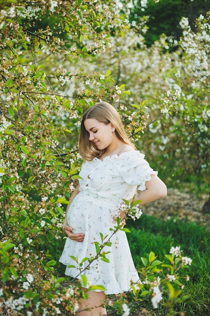 Uma jovem grávida em um vestido branco caminha em um pomar de maçã em flor Mulher grávida feliz Feliz futura mãe