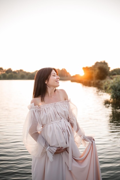 Uma jovem grávida em um vestido delicado à beira do rio com um belo pôr do sol Beleza saúde e maternidade gentil maternidade feliz