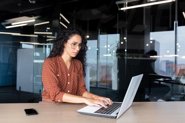Uma jovem hispânica séria trabalhando no escritório em um laptop concentrado digitando no teclado