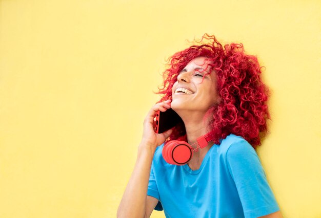 Uma jovem latina com cabelo afro vermelho vestindo uma camiseta azul encostada na parede amarela falando no celular e rindo fones de ouvido vermelhos no pescoço