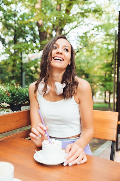 Uma jovem linda com fones de ouvido está sentada em uma mesa em um café de verão e bebendo café ou chá geração z