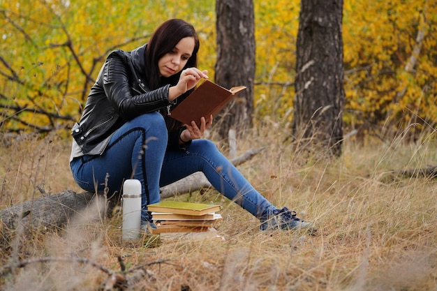 Uma jovem linda com longos cabelos escuros em roupas casuais está sentada em um tronco e lendo um livro na floresta de outono