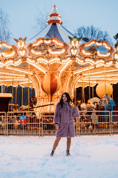 Uma jovem linda com um casaco de pele fica perto do carrossel em uma noite de inverno