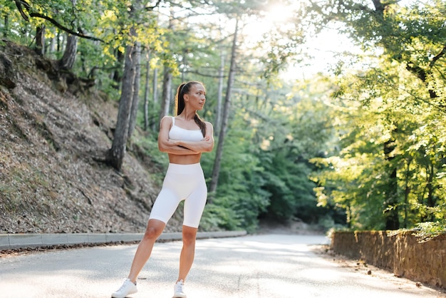 Uma jovem linda em roupas brancas posa antes de um treino de corrida em uma estrada em uma floresta densa durante o pôr do sol Estilo de vida saudável e correndo ao ar livre Juventude