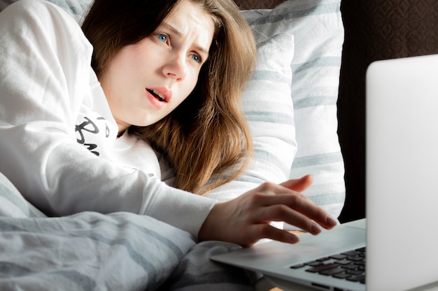 Uma jovem linda em um dia ensolarado em uma jaqueta branca deitada na cama e olhando para o laptop em surpresa