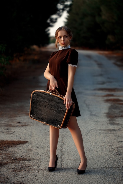 Uma jovem linda em um vestido marrom em estilo retrô está com uma mala preta nas mãos em uma estrada abandonada.