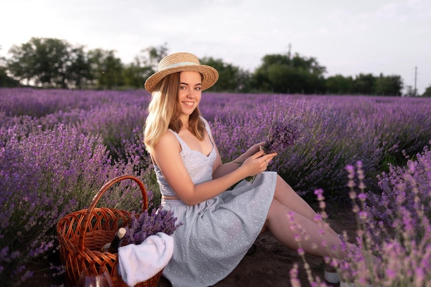 Uma jovem linda sentada entre os arbustos de lavanda segurando um buquê de flores no verão ao pôr do sol. campo de lavanda florescendo e uma senhora de chapéu. Conceito de natureza. acampamento