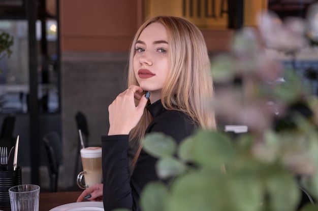 Uma jovem loira atraente e pensativa senta-se à mesa no café e olha para a câmera Retrato de uma jovem no café de verão