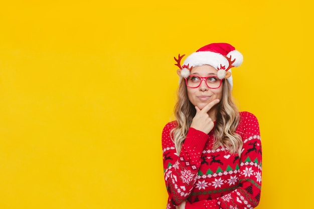 Uma jovem loira com chapéu de Papai Noel de Natal e suéter de cervo vermelho pensa olhando para o espaço da cópia