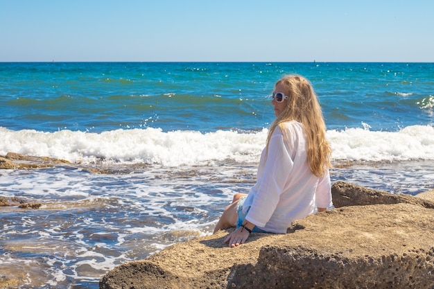 Uma jovem loira com uma túnica branca está sentada em uma pedra à beira-mar com uma onda. viagens e turismo, férias de verão no litoral.