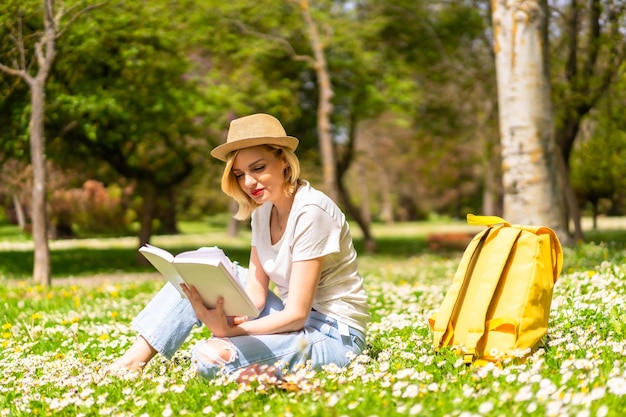 Uma jovem loira de chapéu lendo um livro na primavera em um parque nas férias da cidade ao lado da natureza e ao lado de margaridas sentadas na grama