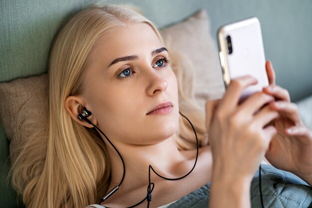 Uma jovem loira está deitada em sua cama sob um cobertor verde. Ela está segurando um telefone ao qual fones de ouvido estão conectados. A garota escolhe qual música ouvir. Ela tem olhos azuis brilhantes