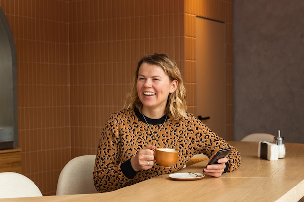 Uma jovem loira está sentada em um café em tons bege bebendo café rolando no telefone e rindo