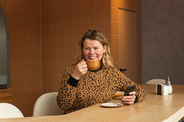 Uma jovem loira está sentada em um café em tons bege bebendo café rolando no telefone e rindo