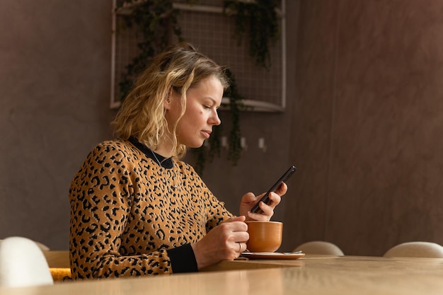 Foto uma jovem loira está sentada em um café em tons bege bebendo café rolando no telefone e sorrindo