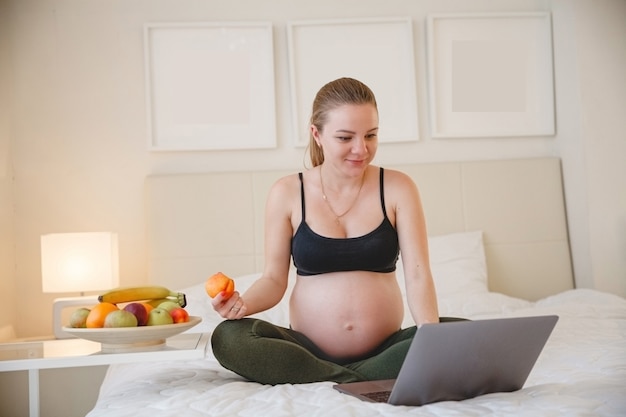 Uma jovem loira grávida está sentada na cama e comendo frutas enquanto olha para seu laptop. Foto de alta qualidade