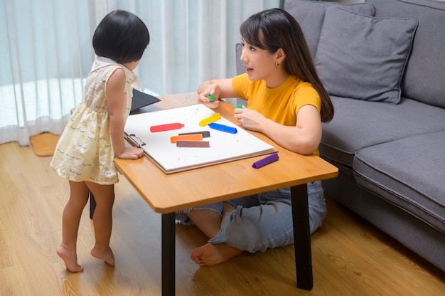 Uma jovem mãe ajudando a filha a desenhar com lápis de cor na sala de estar em casa.