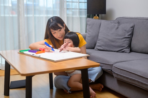 Uma jovem mãe ajudando a filha a desenhar com lápis de cor na sala de estar em casa.