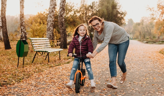 Uma jovem mãe alegre que está ensinando sua filha pequena e sorridente a andar de bicicleta em uma noite ensolarada de outono