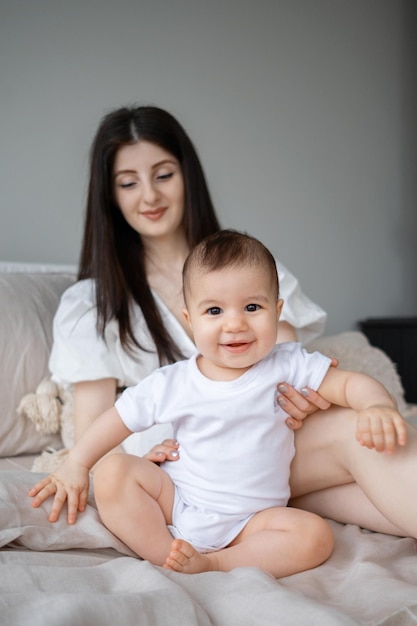 Uma jovem mãe brinca com seu filho sentado na cama Um bebê sorridente está sentado em uma cama com sua mãe em uma casa de campo Uma menina de 9 meses olha para a câmera