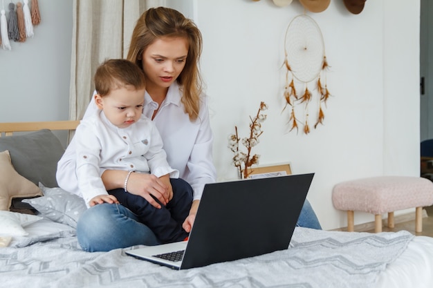 Uma jovem mãe com seu filho amado no quarto na cama com um laptop. Trabalhe em casa com um conceito infantil