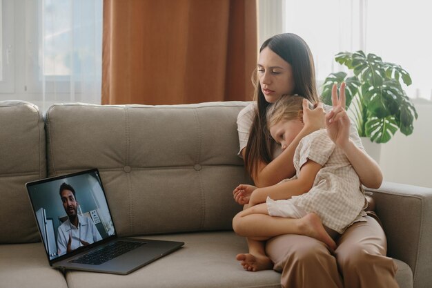 Foto uma jovem mãe com sua filha pequena consulta seu médico de família on-line usando um laptop