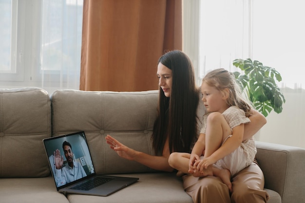 Foto uma jovem mãe com sua filha pequena consulta seu médico de família on-line usando um laptop