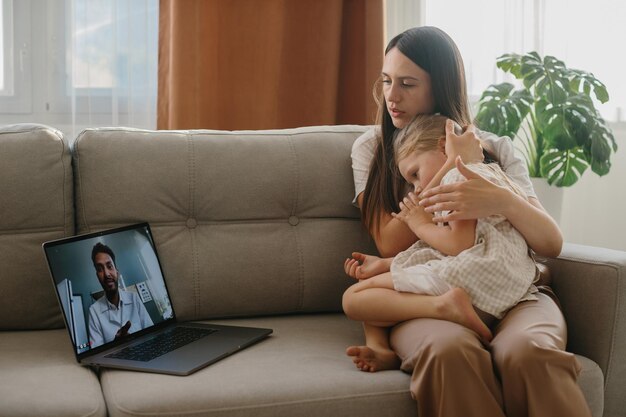 Foto uma jovem mãe com sua filha pequena consulta seu médico de família on-line usando um laptop