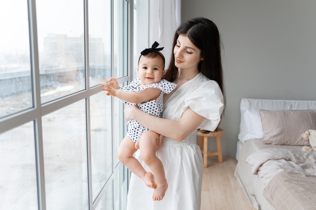 Foto uma jovem mãe com um bebê de 9 meses nos braços fica perto da janela em sua casa de campo