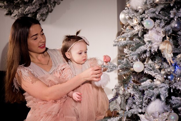 Uma jovem mãe e filha em vestidos combinando decoram a árvore de Natal com bolas novas