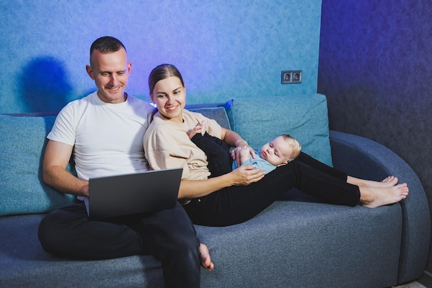 Uma jovem mãe e pai estão sentados com seu filho recém-nascido no sofá e assistindo a um filme em um laptop Relações familiares com um bebê pequeno Um casal de família com uma criança em casa no sofá