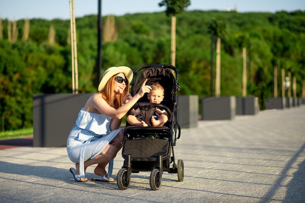 Foto uma jovem mãe feliz caminha com seu bebê em um carrinho no parque no verão sob o sol poente e sorri enquanto se diverte apontando para a distância