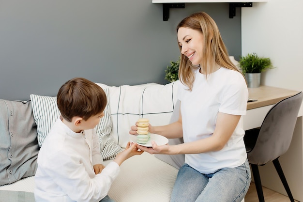 Uma jovem mãe feliz e um filho sorridente em roupas brancas mantêm juntos um prato de bolos de macaroons