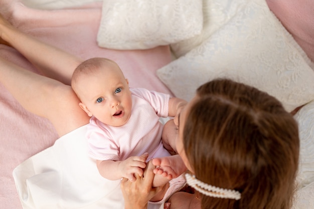 Uma jovem mãe linda segura a filha de uma menina de 6 meses no colo em uma cama branca, brincando e beijando-a