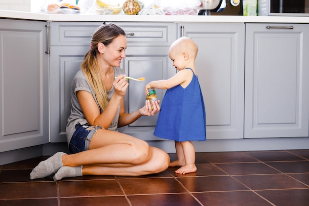 Uma jovem mãe se senta no chão da cozinha e alimenta sua filha de um ano com uma colher.